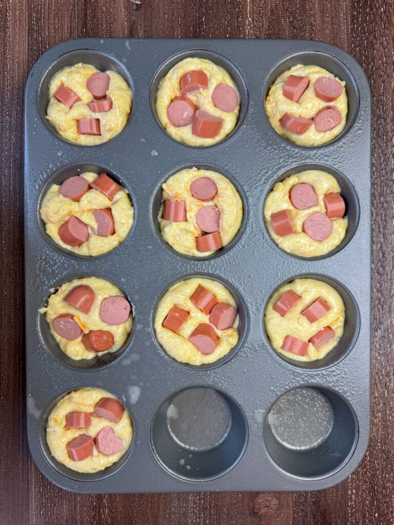 Bite size pieces of hot dogs pressed into muffin tins filled with corn bread mix with shredded cheese.