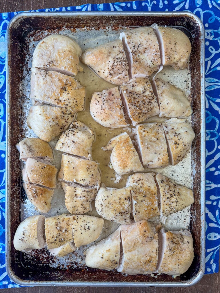Lightly seasoned baked chicken breasts on a large baking sheet cut into thirds and ready to shred.