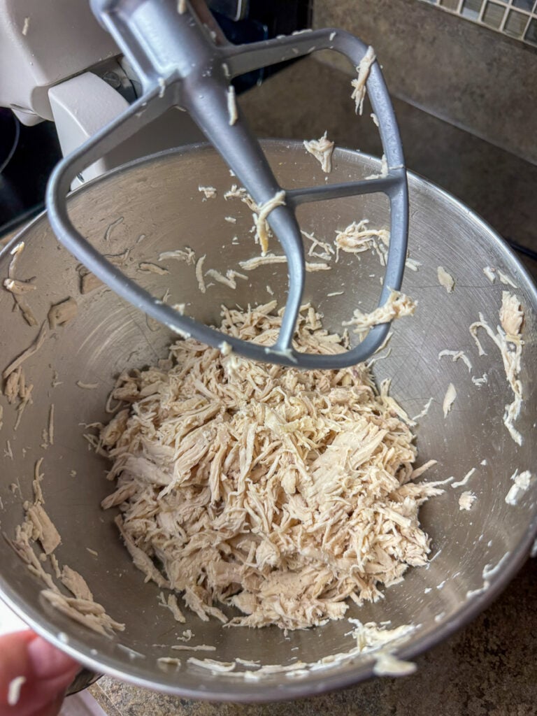 Electric mixer paddle lifted above a silver bowl with finely shredded chicken.
