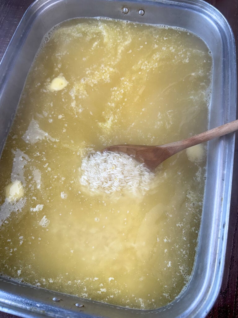 Wooden spoon gently stirring chicken broth into a large roaster of white rice.