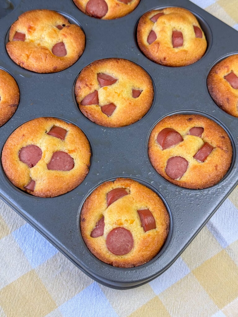 Pieces of hot dog baked info golden brown corn muffins in a muffin tin on a yellow and white plaid napkin.