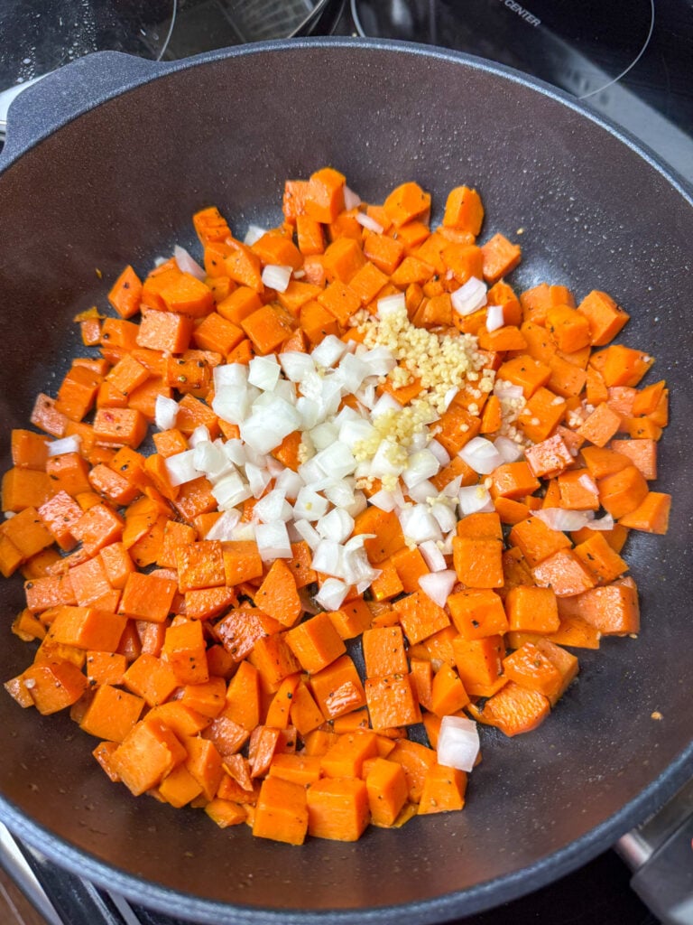 Chopped onion and minced garlic added to a skillet of partially cooked sweet potatoes.