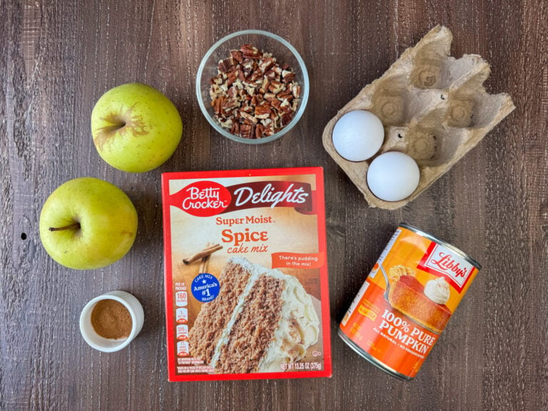 Ingredients to make pumpkin apple cake are apples, pecans, eggs, pure pumpkin, spice cake mix and cinnamon on a wooden counter top.