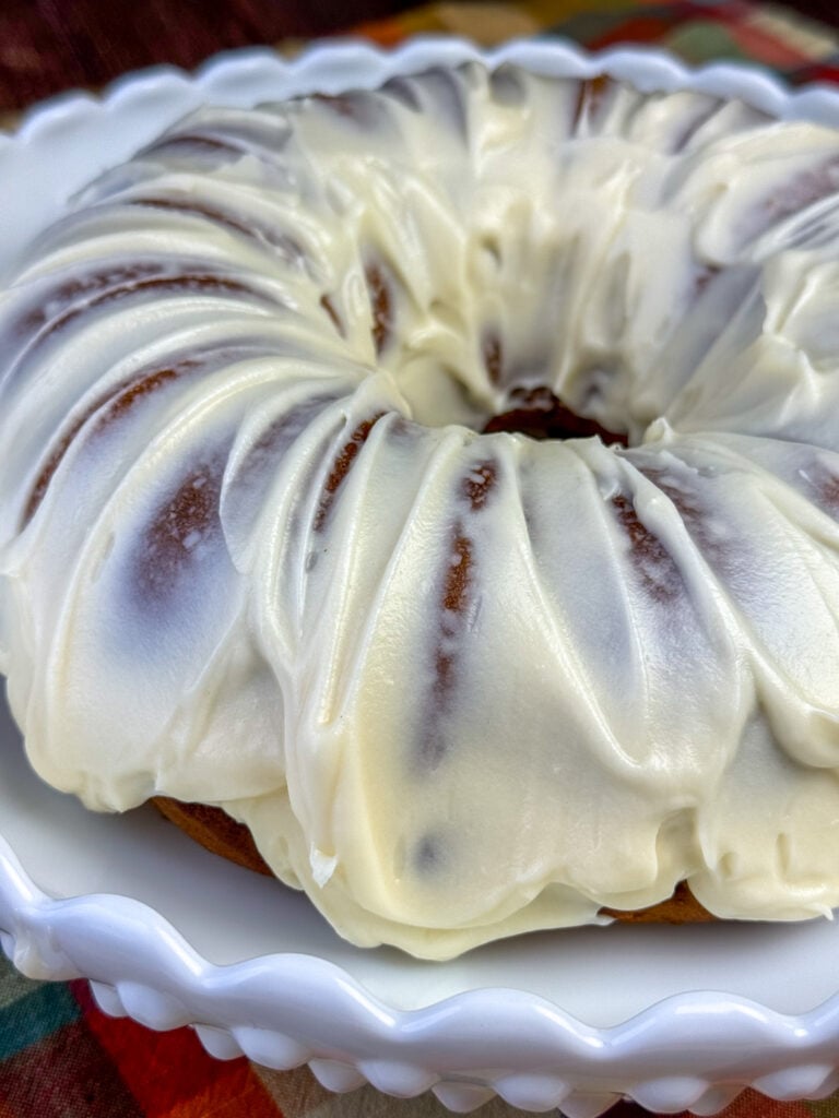 Close up of a pumpkin apple bundt cake coated with cream cheese frosting on a white cake platter.