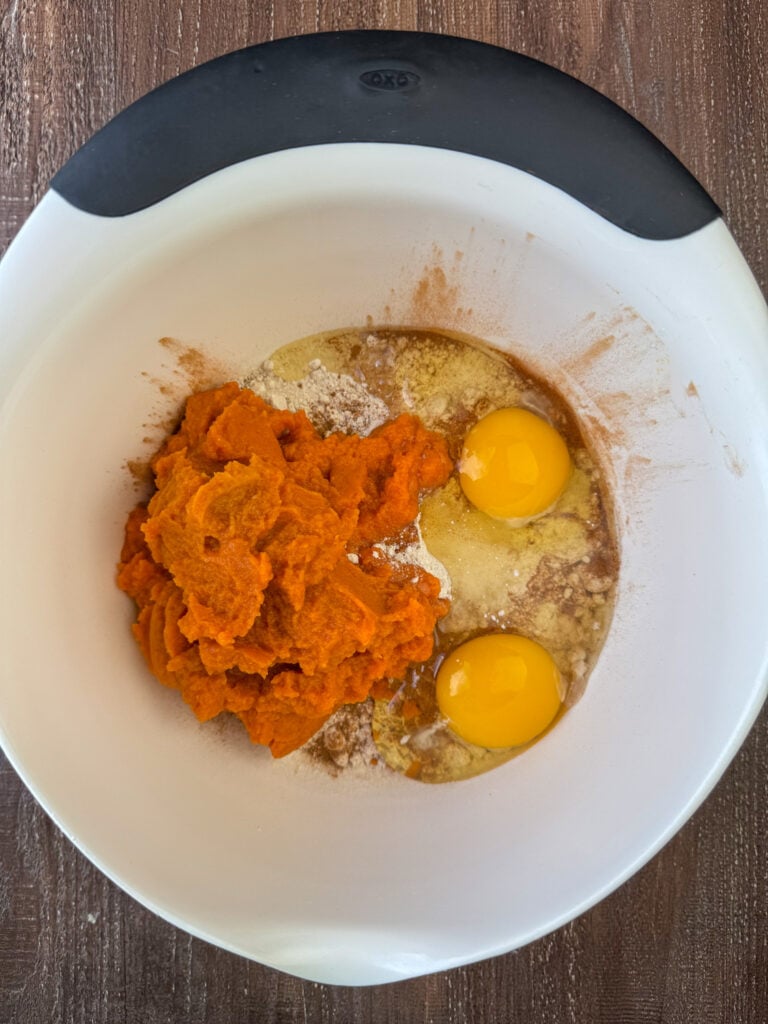 Can of pumpkin puree added to a mixing bowl of spice cake mix, eggs, and cinnamon on a wooden counter top.