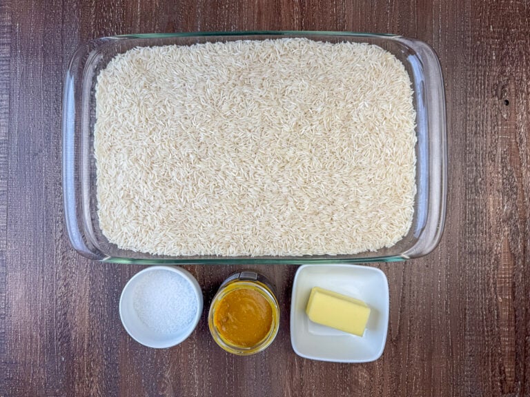 Ingredients to make rice for a crowd are 5 pounds of long grain rice, sea salt, chicken broth base and butter on a wooden counter top.
