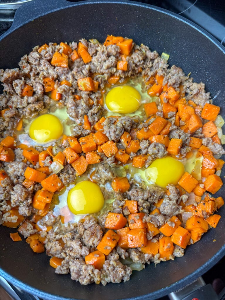 Four whole eggs cracked into a skillet of sausage and sweet potato cubes.