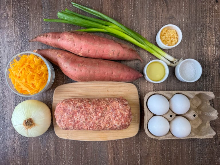 Ingredients to make sausage sweet potato hash are sweet potatoes, green onions, minced garlic, sea salt, olive oil, eggs, Italian sausage, onion and cheddar cheese on a wooden counter top.