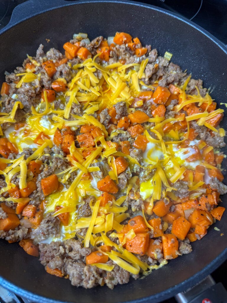 A light layer of cheddar cheese freshly shredded over the sausage and sweet potato hash with eggs in a non-stick skillet.
