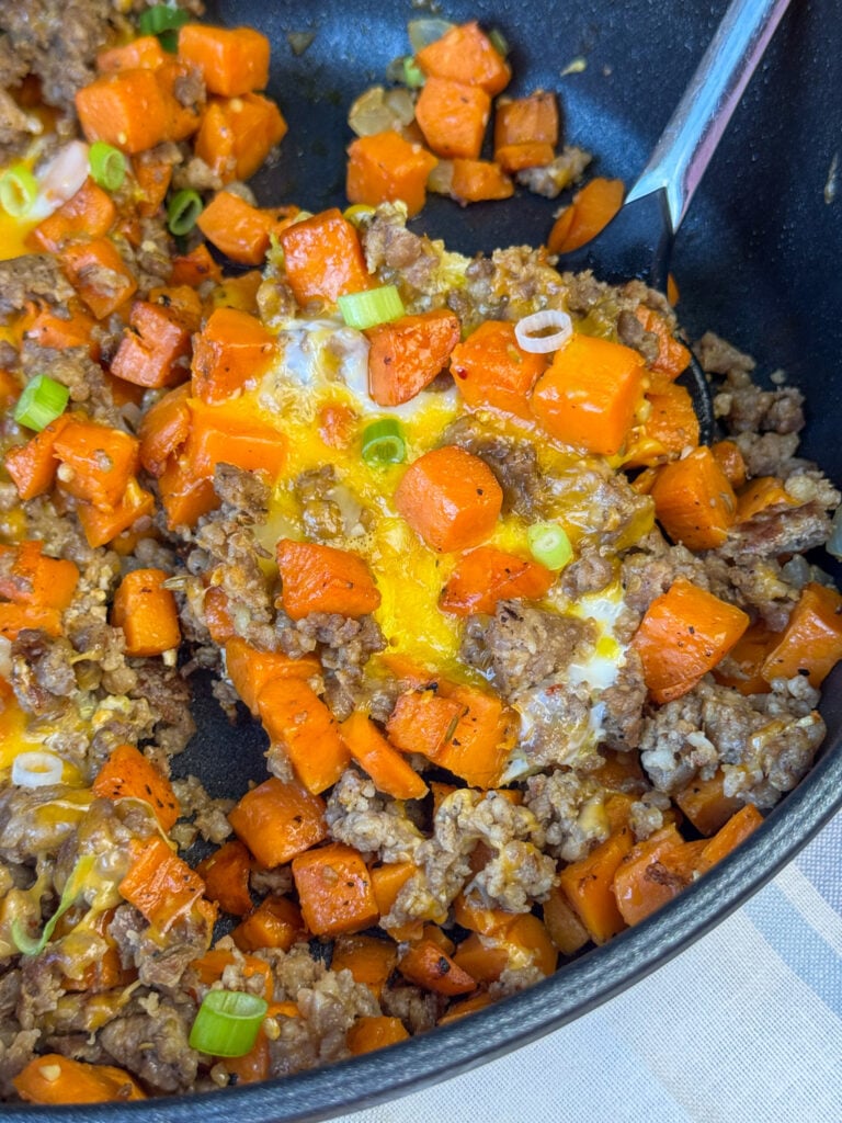 A flavorful mixture of diced sweet potatoes resting on a sausage and egg mixture with a sprinkling of cheddar cheese being lifted on a scoop above the skillet.