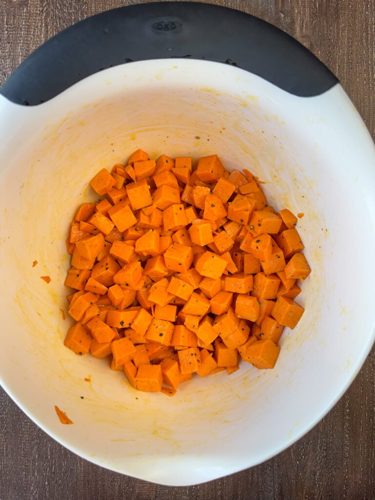 Cubed sweet potatoes coated with olive oil and seasoned with salt and pepper in a white mixing bowl.