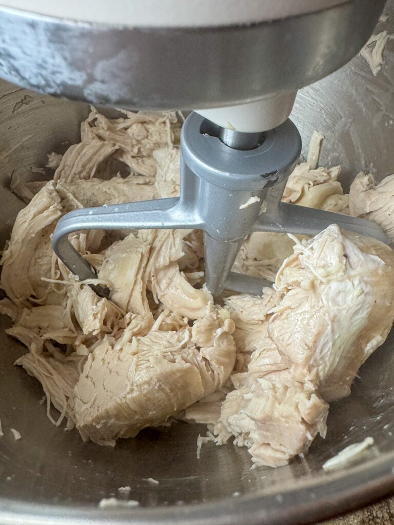Close up chunks of tender chicken breasts in a mixer with the paddle ready to spin and shred them.