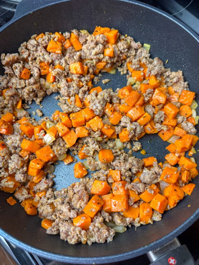 Cooked and crumbled sausage and cubes of sweet potatoes in a skillet with four wells made to add eggs to.