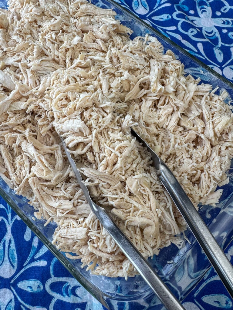 Glass serving dish full of warm, shredded chicken for a crowd with tongs ready to serve.