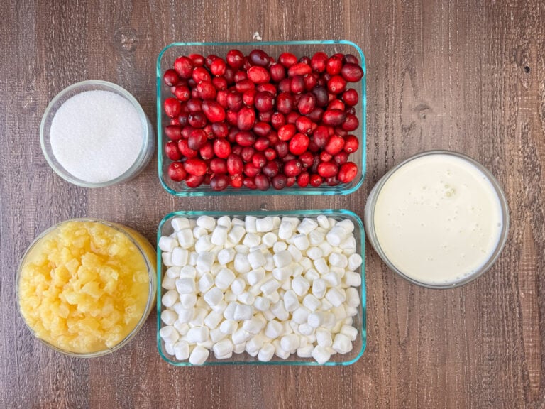 Ingredients to make cranberry fluff are whole cranberries, mini marshmallows, crushed pineapple, sugar and whipping cream on a wooden counter top.