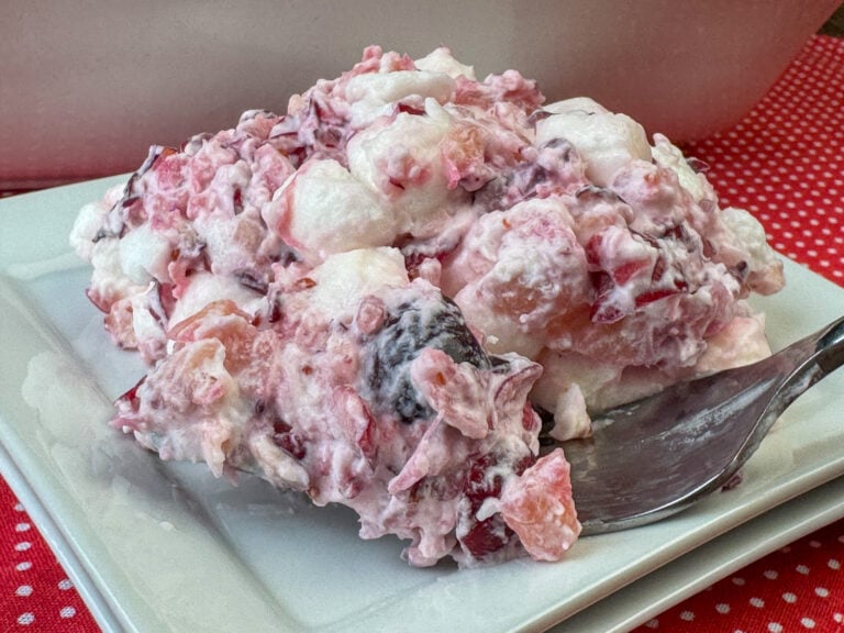 Close up of a fork ready to lift a bite of whipped cream coated bits of cranberries, pineapple and mini marshmallows on a small white plate.