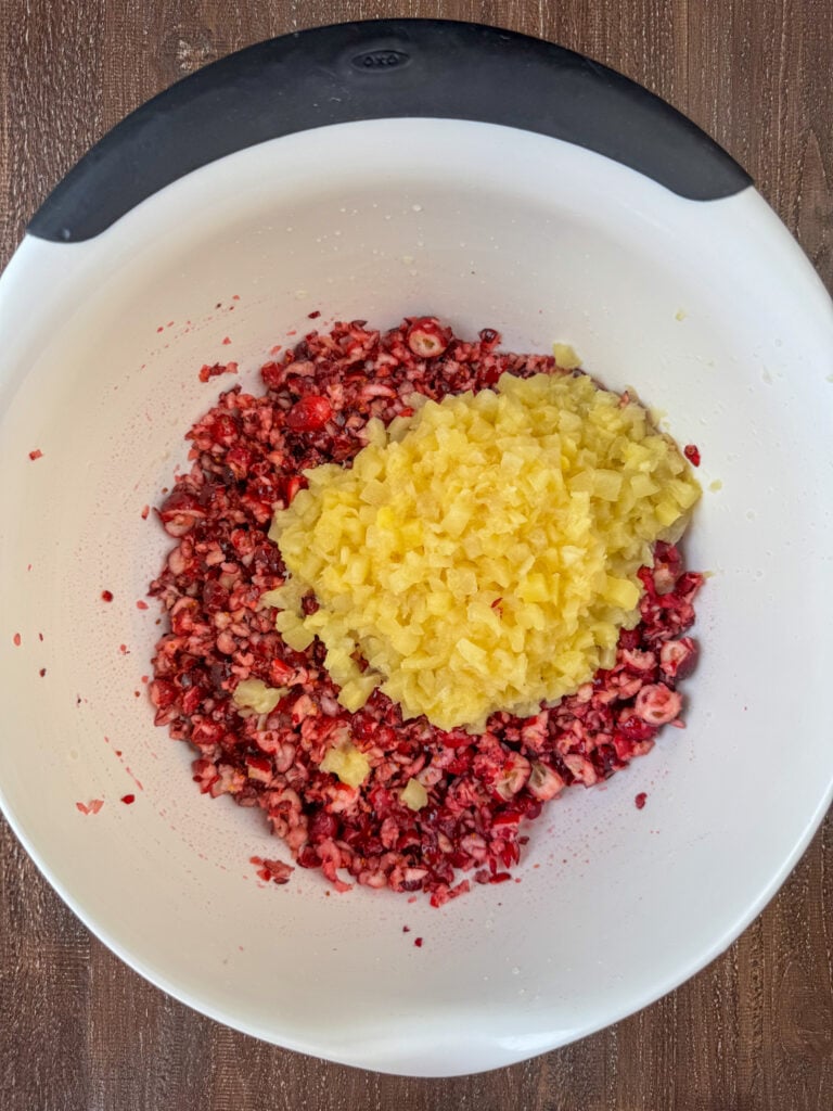 Drained crushed pineapple added to chopped cranberries in a white bowl.