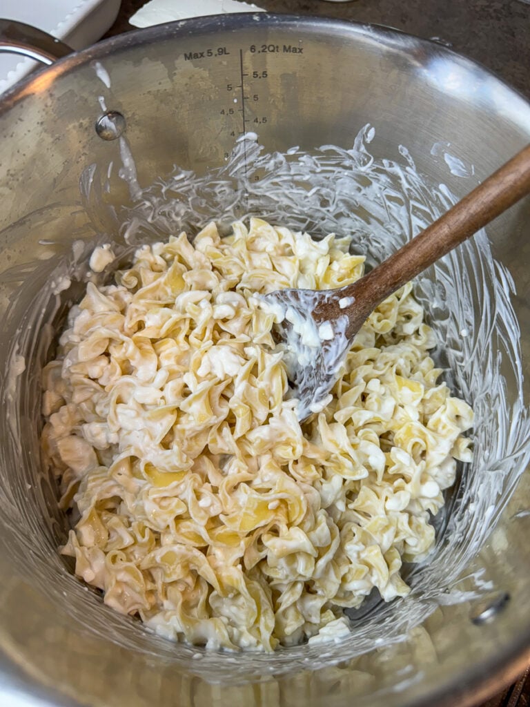 Wooden spoon stirring sour cream and cottage cheese into warm egg noodles in a post.