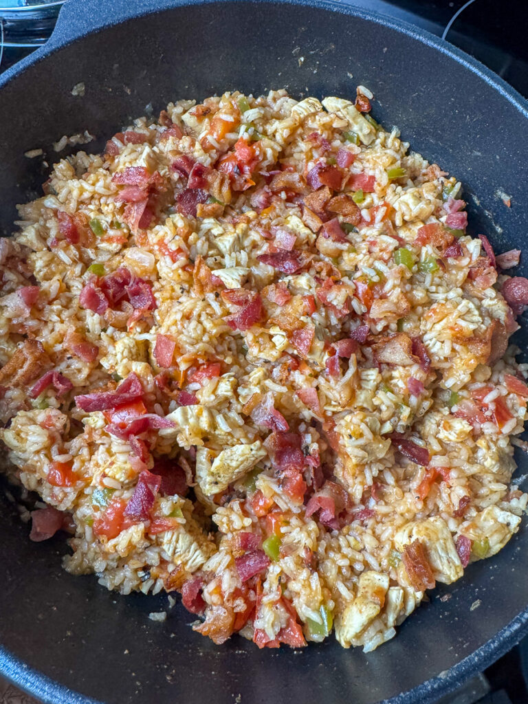 Pieces of chicken breast and bacon stirred into rice, onion, and green peppers in a skillet.
