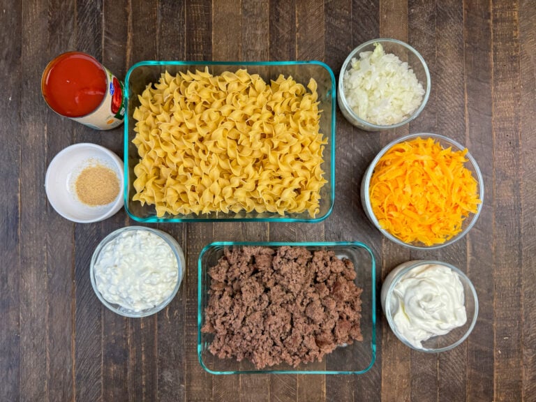 Ingredients to make sour cream noodle bake are egg noodles, onions, cheddar cheese, sour cream, ground beef, cottage cheese, garlic powder and tomato sauce on a wooden counter top.