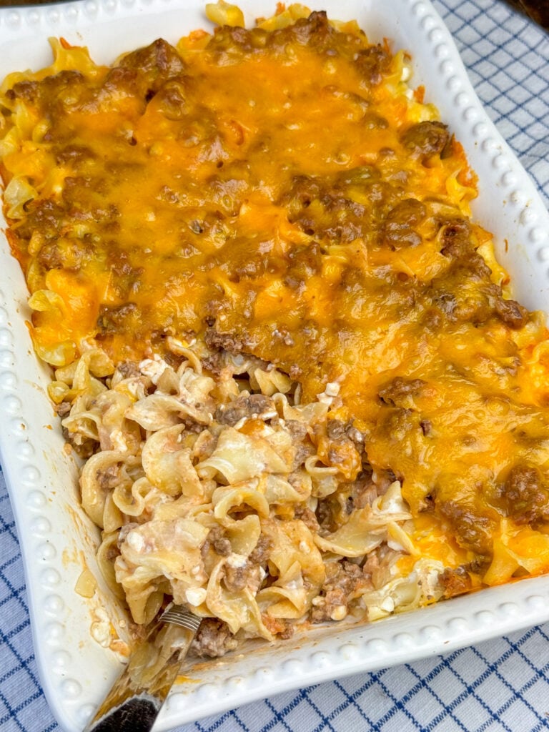 Serving spoon scooping down the corner of a sour cream noodle bake with a thick layer of cheddar cheese on top.